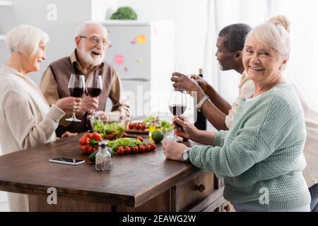 happy senior woman holding glass of red wine and smiling near multicultural retired friends on blurred background Stock Photo
