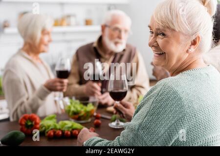 happy senior woman holding glass of red wine near retired friends on blurred background Stock Photo