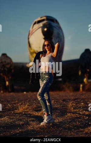 A tall blonde teenage girl wearing  ripped jeans and a white tank top poses for portraits by abandoned airplanes covered in graffiti in the desert wild Stock Photo