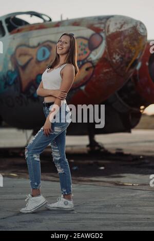 A tall blonde teenage girl wearing  ripped jeans and a white tank top poses for portraits by abandoned airplanes covered in graffiti in the desert wild Stock Photo