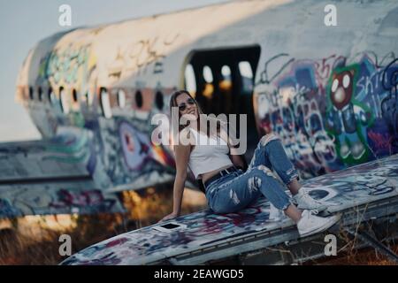 A tall blonde teenage girl wearing  ripped jeans and a white tank top poses for portraits by abandoned airplanes covered in graffiti in the desert wild Stock Photo