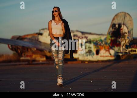 A tall blonde teenage girl wearing  ripped jeans and a white tank top poses for portraits by abandoned airplanes covered in graffiti in the desert wild Stock Photo