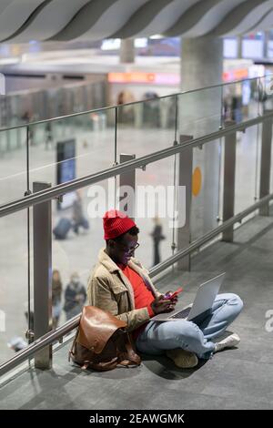 African American traveler millennial man resting and sitting on the floor in airport using cellphone Stock Photo