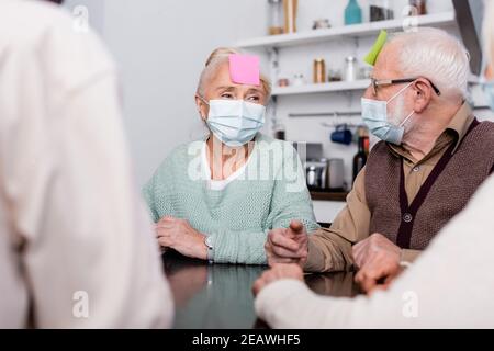 senior man and woman in medical masks with sticky notes on foreheads playing game with multicultural friends on blurred foreground Stock Photo