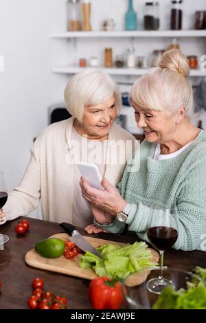 happy senior woman holding smartphone near retired friend in kitchen Stock Photo