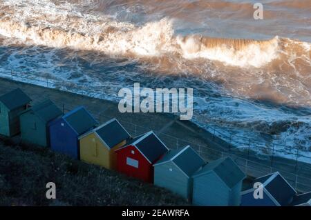 Huge crashing waves break on a beach next to a row of beach huts Stock Photo