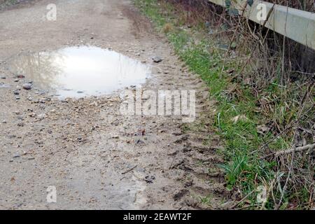 Country road with a pothole filled with rain water. Tyre traces and imprints are visible in soft, muddy surface of the road. Stock Photo
