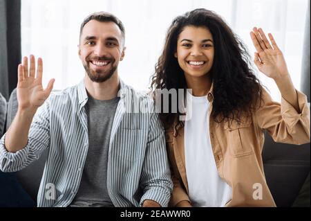 Two colleagues, students or couple in love, caucasian guy and african american girl, sitting on a sofa talk on video call with colleagues or friends g Stock Photo