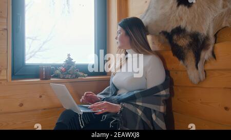 Lonely young caucasian woman sitting near the window covered with blanket lookin outside and using laptop. High quality photo Stock Photo