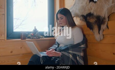 Young caucasian woman sitting near the window covered with blanket and using laptop. High quality photo Stock Photo
