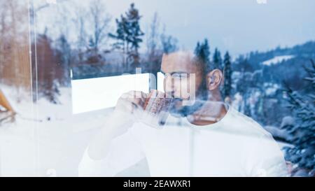 Portrait of young man drinking hot beverage wile looking through the window at first snow. Christmas celebration. High quality photo Stock Photo