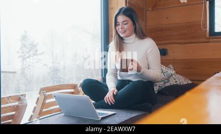 Young caucasian woman drinking coffee and using laptop near the window in winter. High quality photo Stock Photo
