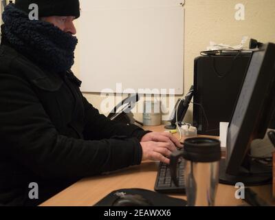 Keeping warm, working in a cold office, man working at desk wearing a thick winter coat, hat and scarf Stock Photo