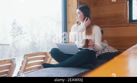 Young woman drinking coffee near the window and using laptop for video call. High quality photo Stock Photo