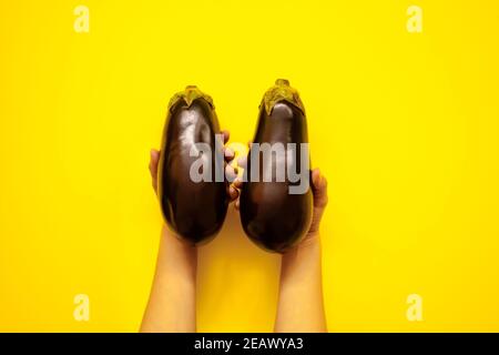 Top view of woman hands holding a pair of aubergines in studio over a yellow background - flatlay look Stock Photo