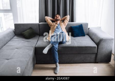 Satisfied handsome caucasian man is sitting on the couch in living roo, thinking about plans for weekends or vacation and smile. Young freelancer rest Stock Photo