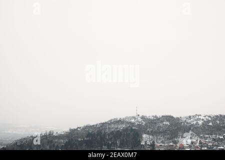 Minimalist view with a lot of negative space of snow covered rocky hill with trees and TV antenna at the top, rising above small group of houses Stock Photo