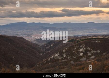 View over a canyon with cliffs covered by autumn colored trees and distant misty cityscape under an orange, cloudy, sunset sky Stock Photo