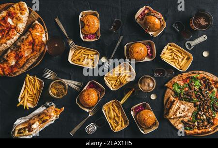 Lockdown takeaway fast food dinner from delivery service concept. Flat-lay of quarantine home party with burgers, french fries, sandwiches, pizza, beer in glasses over dark table, top view Stock Photo