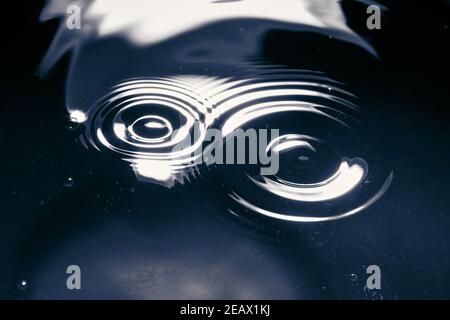 Water droplets falling on water causing waves in circles on a blue background with white reflections. Stock Photo