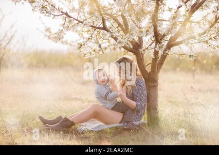 Young curly-haired mother hugs little son, sitting in the green grass near blooming white cherry tree. Family walk in the garden. Weekend rest outside Stock Photo