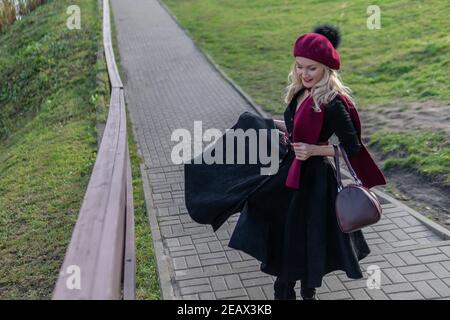 A joyful woman walks up the stairs in a burgundy palette and biret, with beautiful eyes in black clothes, in the fall against the background of a pond Stock Photo