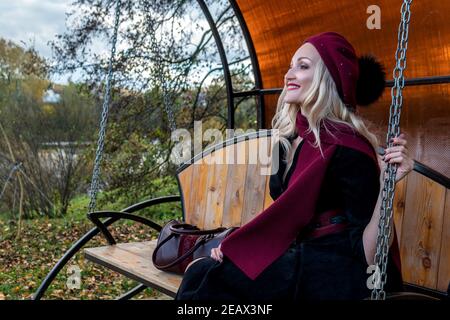 A cheerful woman sits on a garden swing, forged in a burgundy coat and biret, smiles at the camera, in the fall against the background of trees, a blu Stock Photo