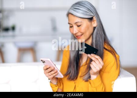 Defocused senior gray-haired woman is shopping online. Joyful asian gray-haired woman uses the credit card and mobile phone for shopping and payment o Stock Photo