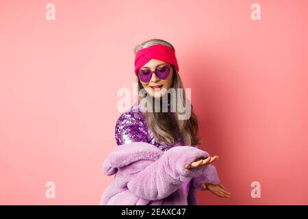 Fashionable mature asian woman in glittering dress dancing on party, having fun and relaxing, standing over pink background Stock Photo