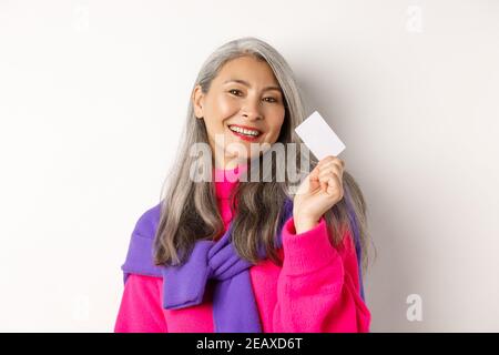 Shopping concept. Stylish asian senior woman smiling and showing plastic credit card, paying contactless, standing over white background Stock Photo