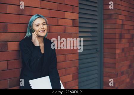 Delightful caucasian businesswoman with blue hair discussing on phone and looking at camera while posing on a brick wall with a computer Stock Photo