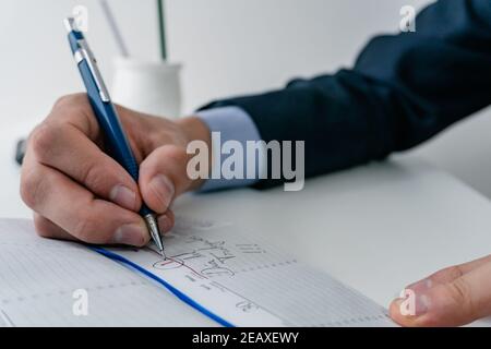 young businessman writing with a mechanical pencil in his notebook may 1 labor day. wearing blue suit, with computer and smart phone by his side. Stock Photo