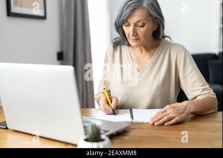 Mature focused woman watching online webinars on the laptop sitting at the table at home, taking notes, e-learning. Concentrated senior business woman Stock Photo