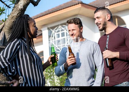 Gay couple and African American woman friend enjoying chatting and drinking alcohol outdoors at the house on weekend Stock Photo