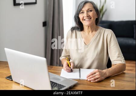 Charming mature mid-aged woman watching online webinars on the laptop sitting at the table at home, taking notes, e-learning. Senior business lady Stock Photo