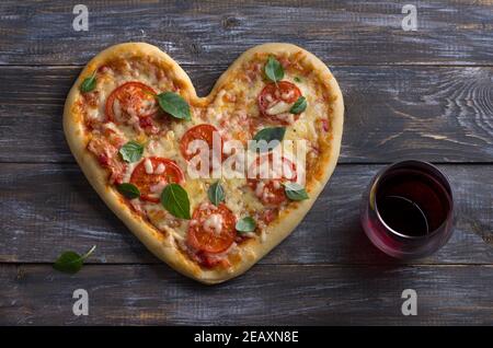 Homemade delicious margarita pizza with tomatoes and basil in the shape of a heart with a glass of red wine on a wooden table for Valentine's Day, top Stock Photo