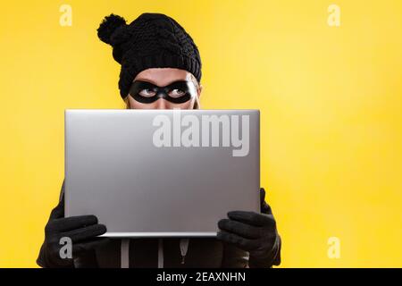 The concept of cybercrime and hacking. Portrait of a woman in a black hat, gloves and mask, who holds a laptop in her hands, covering her face with it Stock Photo