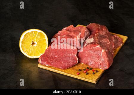 Fresh raw veal tenderloin meat in large chunks and sliced medallions with lemon and pepper peas on a wooden chopping board on a dark vintage metal bac Stock Photo