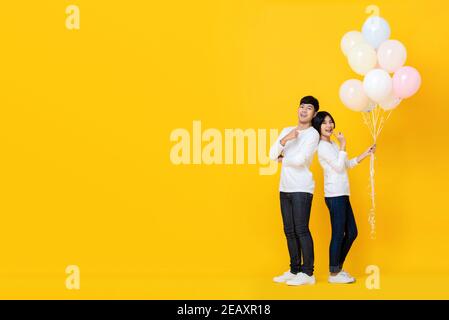 Cheerful young Asian couple standing with back to back support holding bunch of balloons against colorful yellow background Stock Photo