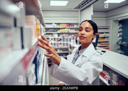 Young woman working in pharmacy looking for medicine in shelf standing behind counter Stock Photo