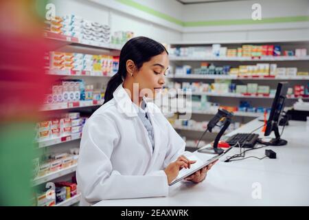 Young female pharmacist standing behind counter wearing labcoat using digital tablet in chemist Stock Photo