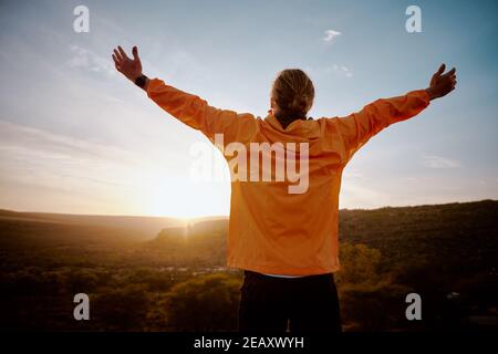 Rear view of young man standing on mountain with outstretched arms feeling relaxed after morning running in nature Stock Photo