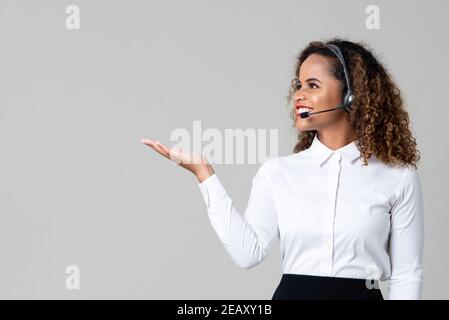 Smiling service minded African American woman wearing headphones as a call center staff with hand open on light gray background Stock Photo