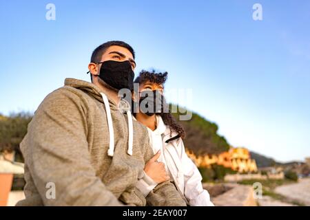 MIxed-race afro-american girl embracing her Caucasian boyfriend sitting outdoor in sea resort looking the sunset wearing black protective mask against Stock Photo
