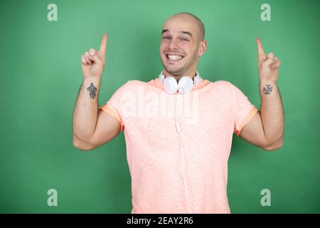 Young bald man using headphones over green background smiling, looking at the camera and pointing up with fingers and raised arms Stock Photo