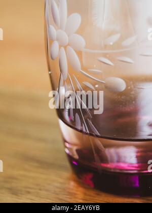 A vintage drinking glass, etched flower design, pinky red purple wine coloured tinted, with a small amount of water in it on a wooden table Stock Photo