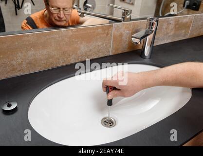 The plumber with a screwdriver fixes the sink drain in the bathroom. Stock Photo