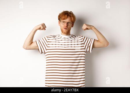 Image of confident and strong redhead man flexing biceps, showing muscles after gym, standing over white background Stock Photo