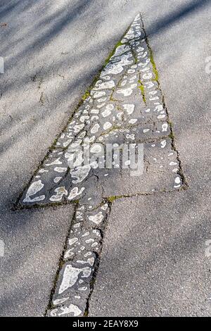 Road markings arrow with cracked peeled white paint on an old road with cracks and overgrown grass in the shade of trees Stock Photo
