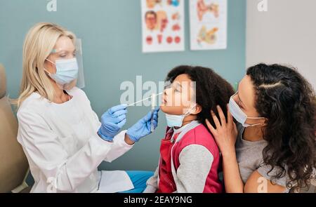 African American female kid having PCR test during coronavirus epidemic. doctor wearing a protective shield using a cotton swab makes a kid's nasal ca Stock Photo